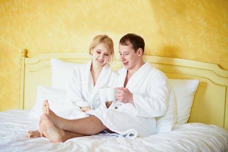 Straight couple in white bathrobes drinking coffee in bed