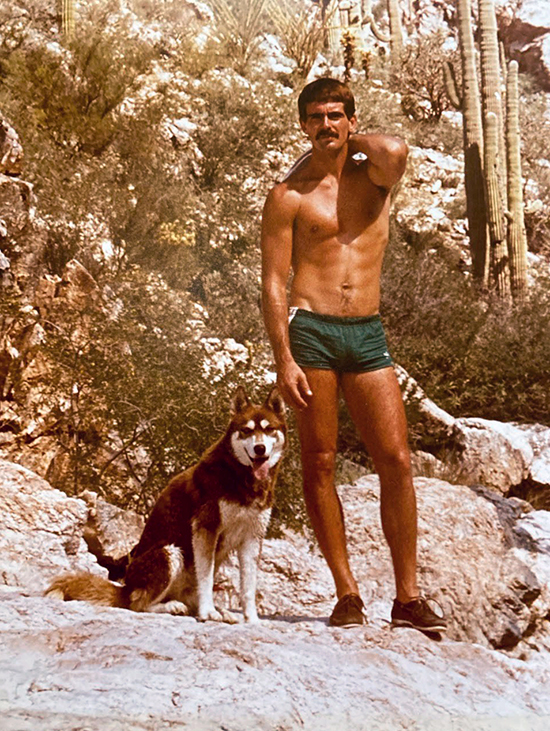Will seagers, shirtless and wearing green shorts, in the desert beside a cactus with a dog beside him, late 1970s